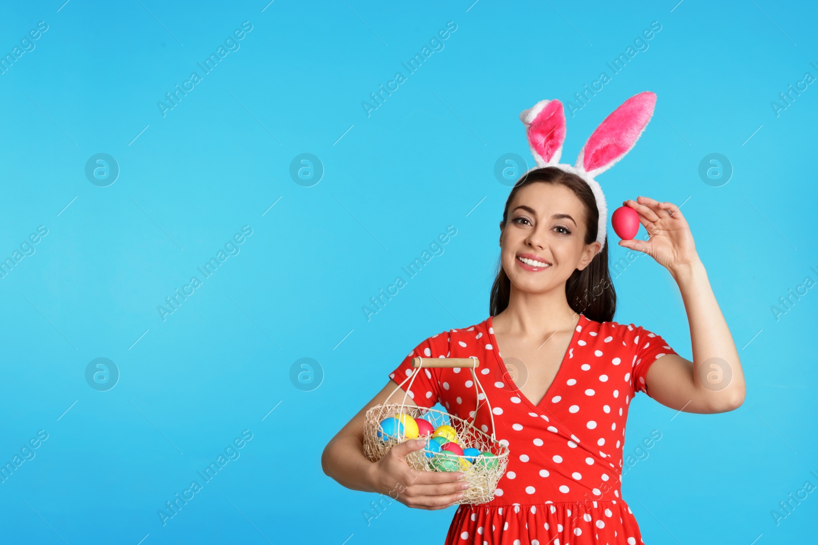 Photo of Beautiful woman in bunny ears headband holding basket with Easter eggs on color background, space for text