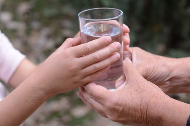 Photo of Child giving glass of water to elderly woman outdoors, closeup