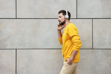 Handsome man with headphones walking near grey stone wall outdoors, space for text