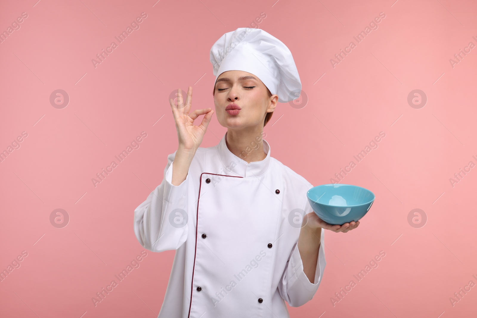 Photo of Young woman chef in uniform holding bowl and showing perfect sign on pink background