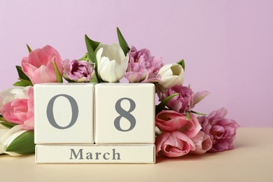 Wooden block calendar with date 8th of March and tulips on table against lilac background. International Women's Day