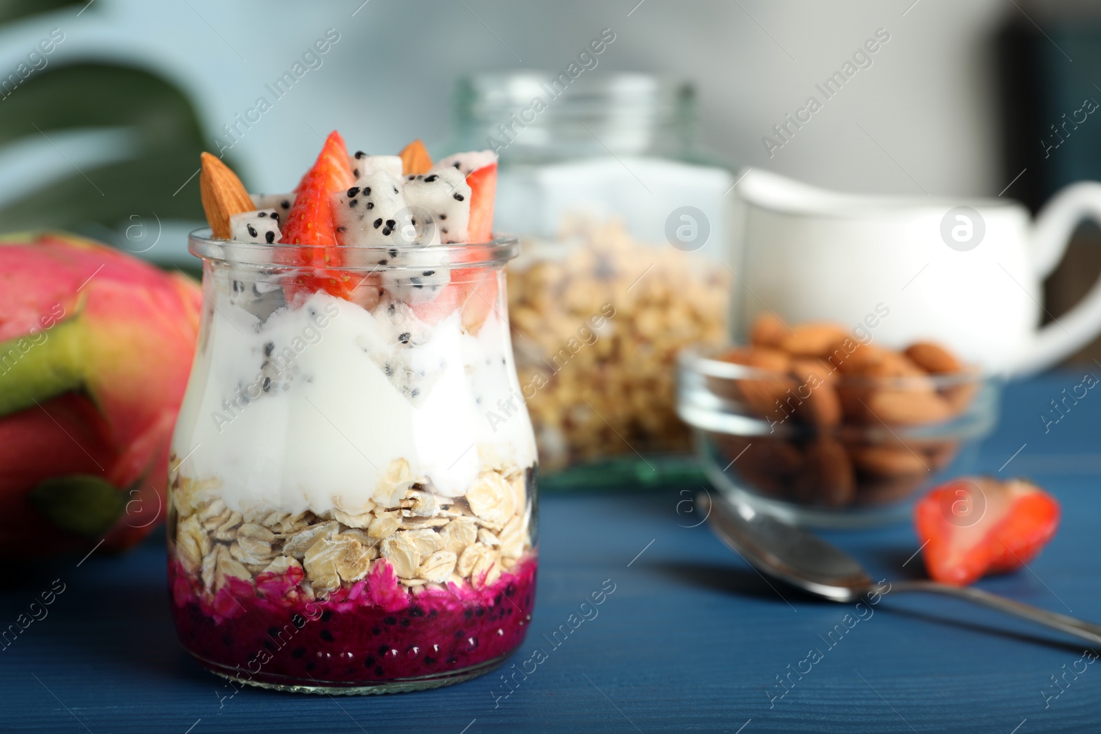 Photo of Glass jar of granola with pitahaya, yogurt and strawberries on blue wooden table, space for text