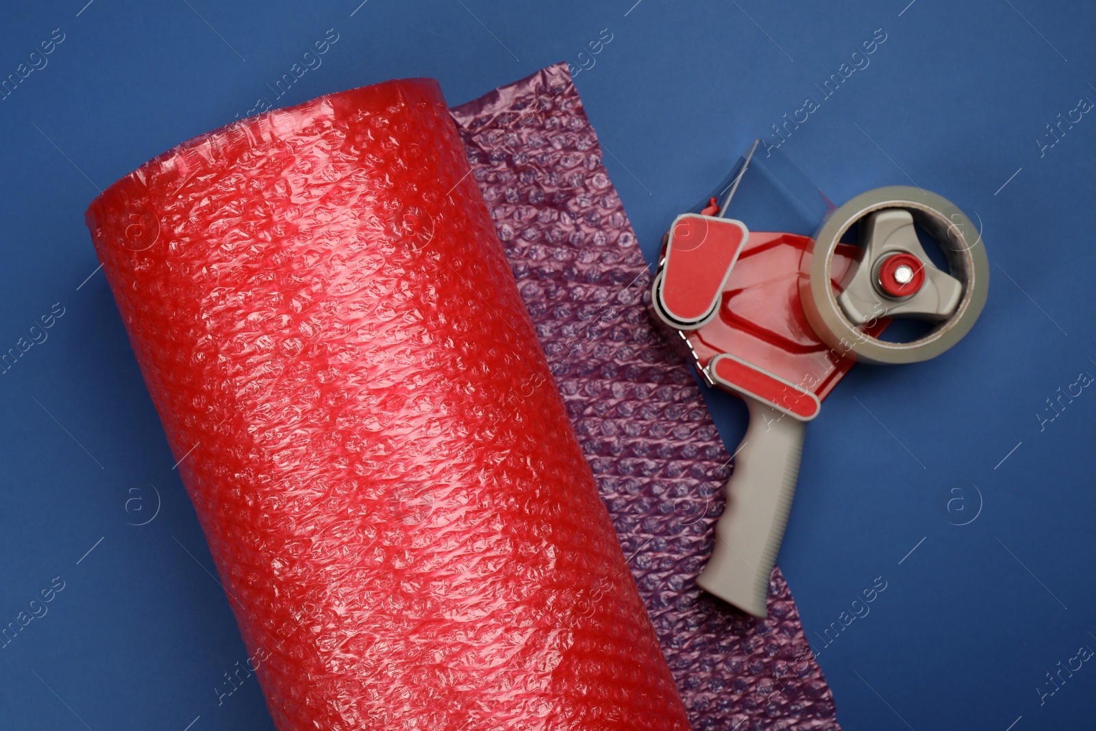 Photo of Bubble wrap roll and tape dispenser on blue background, flat lay