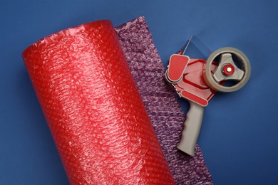 Photo of Bubble wrap roll and tape dispenser on blue background, flat lay