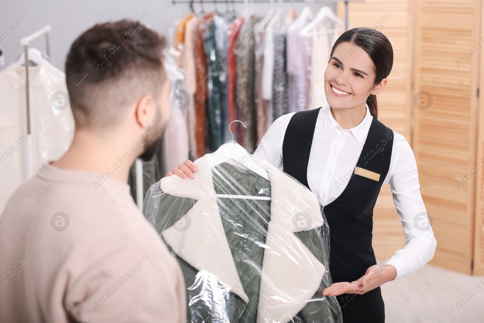 Photo of Dry-cleaning service. Happy worker giving coat in plastic bag to client indoors