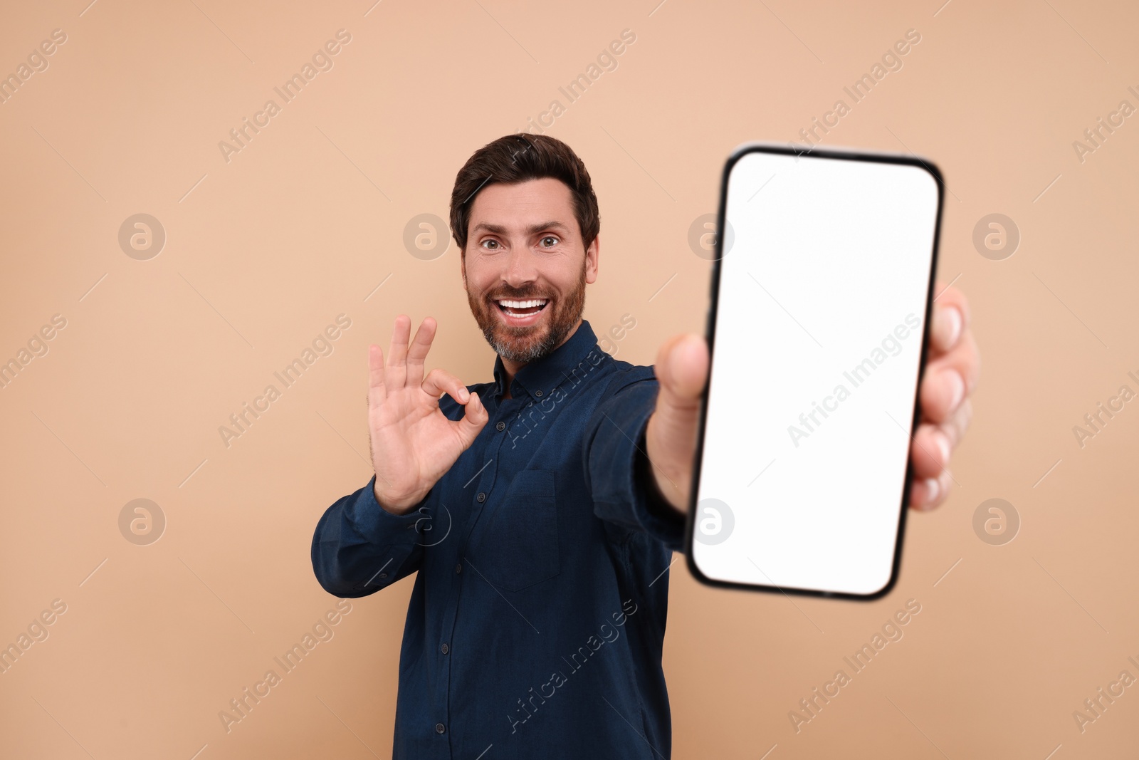 Photo of Handsome man showing smartphone in hand and OK gesture on light brown background