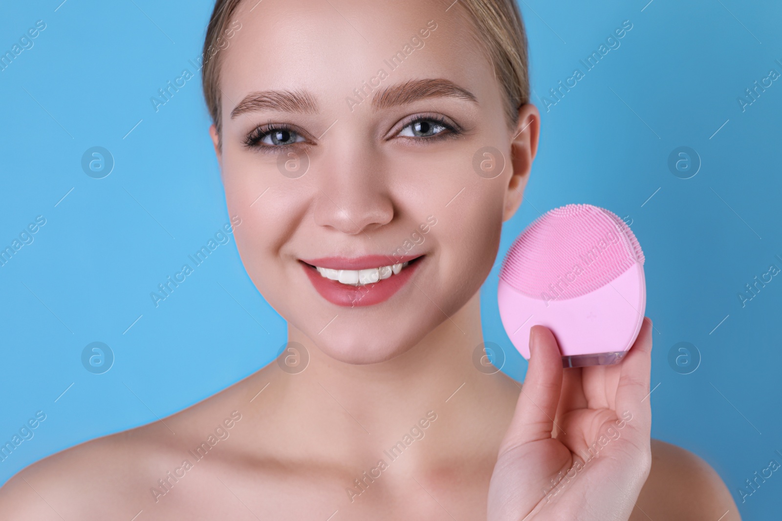 Photo of Young woman washing face with cleansing brush on light blue background. Cosmetic product