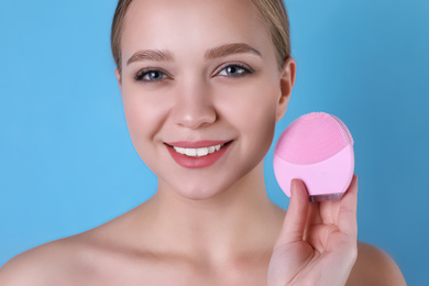 Photo of Young woman washing face with cleansing brush on light blue background. Cosmetic product
