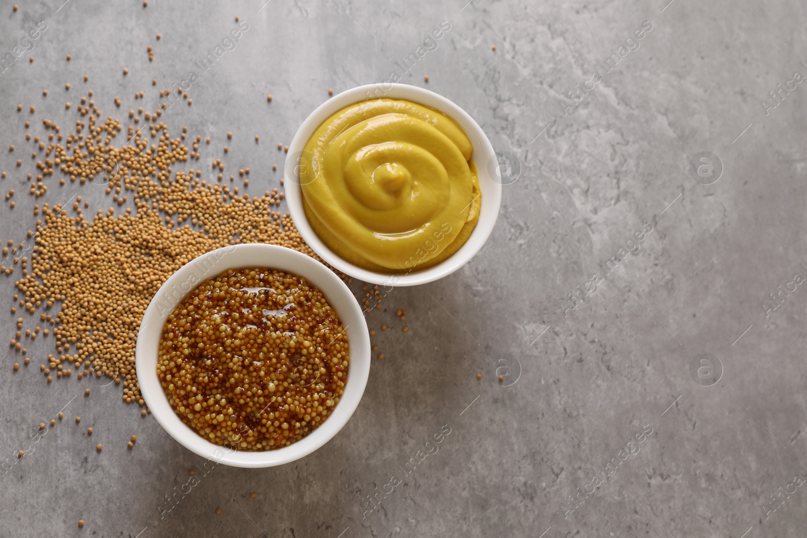 Photo of Bowls with mustard and seeds on grey table, flat lay. Space for text