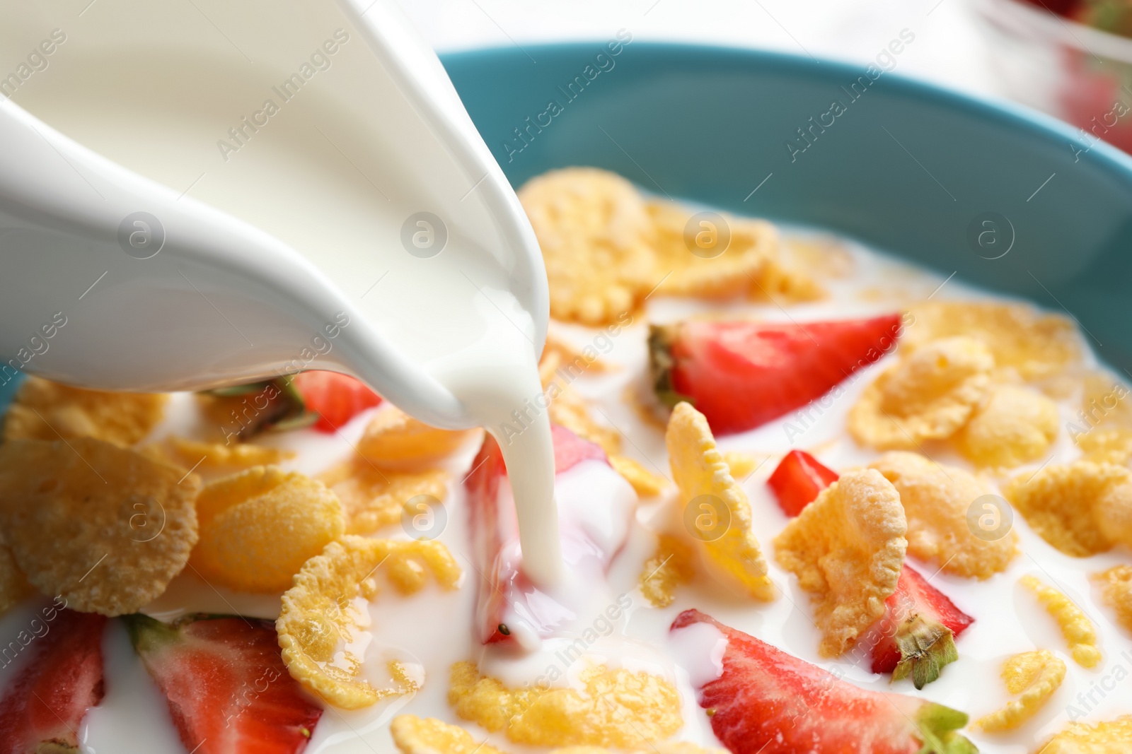 Photo of Pouring milk into bowl with crispy corn flakes and strawberries, closeup