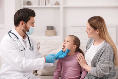 Photo of Doctor in medical mask examining girl`s oral cavity with tongue depressor near her mother at home
