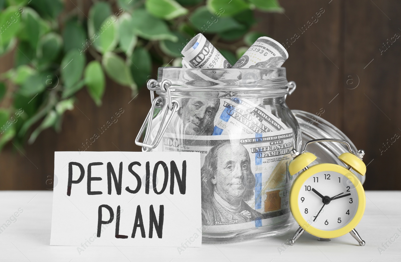 Photo of Card with phrase Pension Plan, dollar banknotes in glass jar and alarm clock on white wooden table. Retirement concept