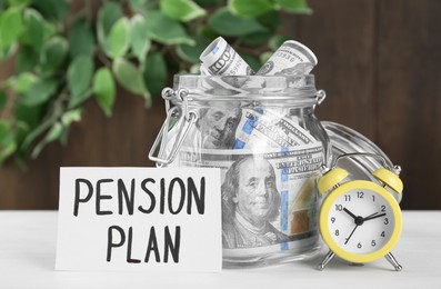 Card with phrase Pension Plan, dollar banknotes in glass jar and alarm clock on white wooden table. Retirement concept