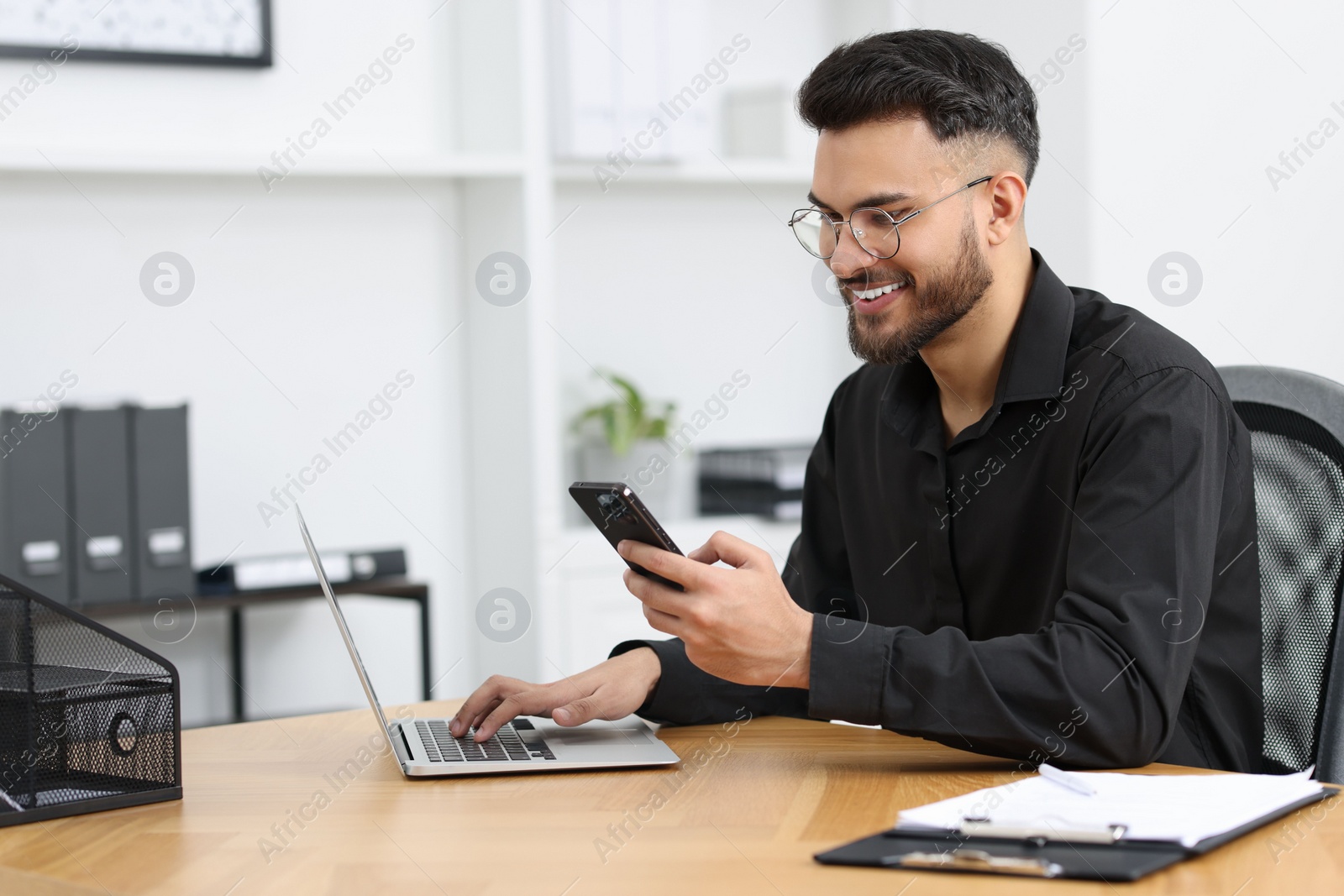 Photo of Handsome young man using smartphone while working with laptop at wooden table in office