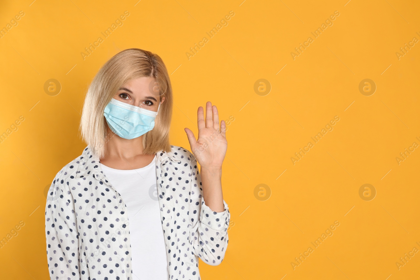 Photo of Woman in protective mask showing hello gesture on yellow background, space for text. Keeping social distance during coronavirus pandemic