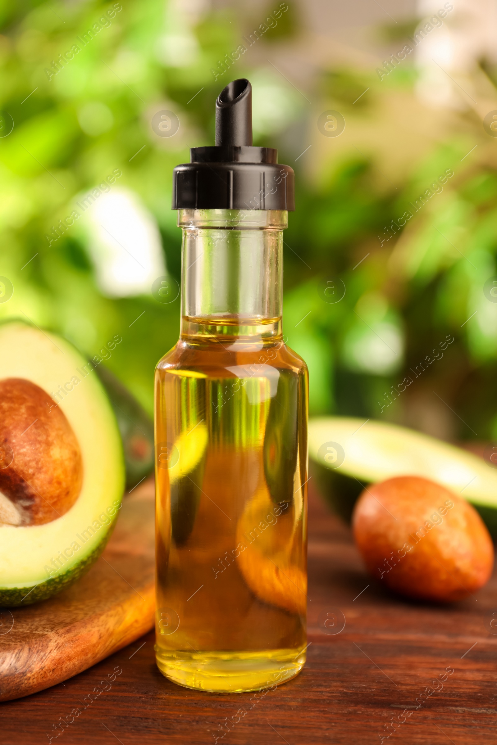 Photo of Glass bottle of cooking oil and fresh avocados on wooden table against blurred green background