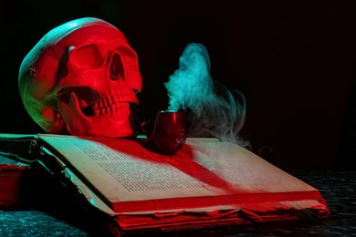 Photo of Human skull with smoking pipe and old book in neon lights on black background