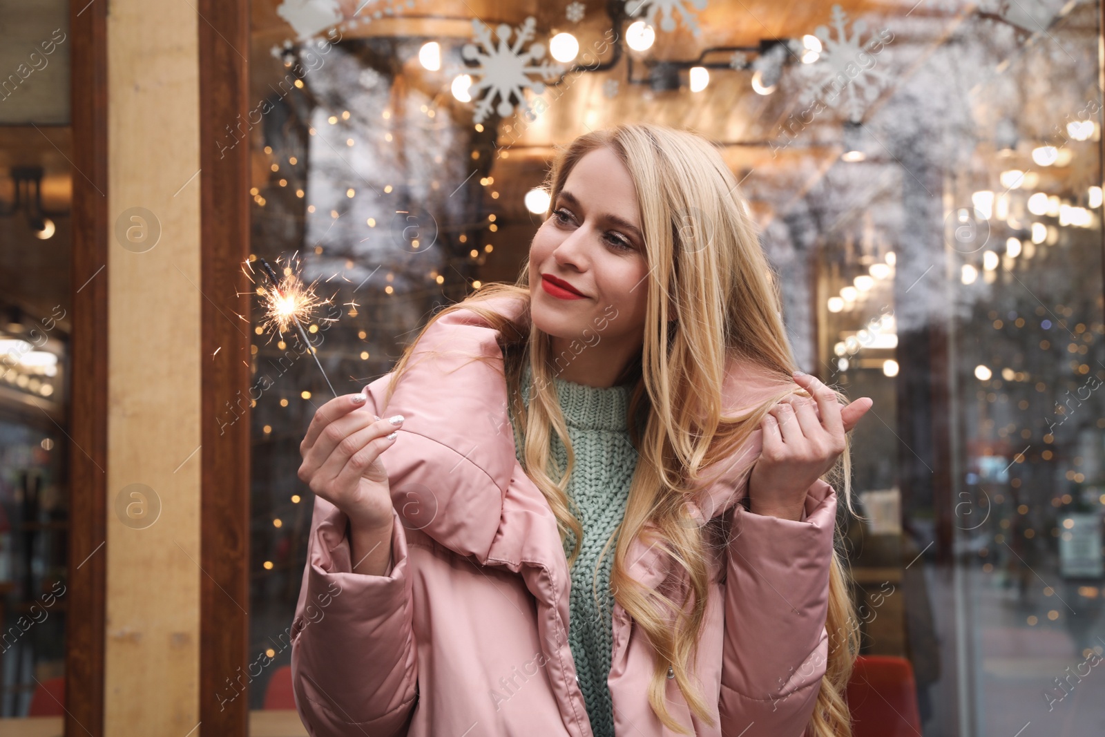 Photo of Young woman with burning sparkler near cafe decorated for Christmas