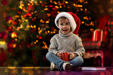 Photo of Little boy in Santa Claus cap with drink near Christmas tree at home