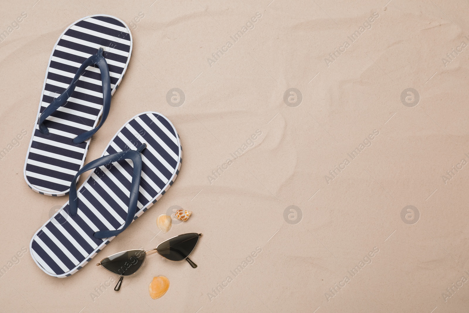 Photo of Pair of striped flip flops, sunglasses and sea shells on sand, flat lay. Space for text