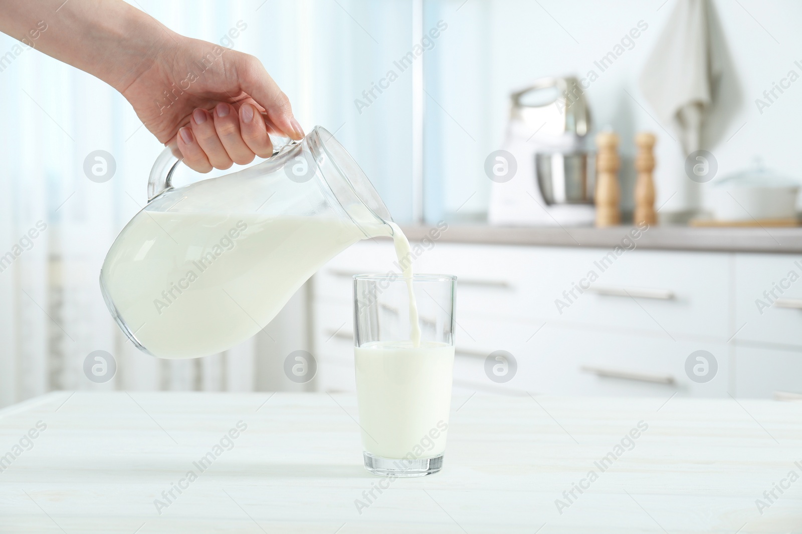 Photo of Woman pouring milk from jug into glass at white wooden table in kitchen, closeup. Space for text