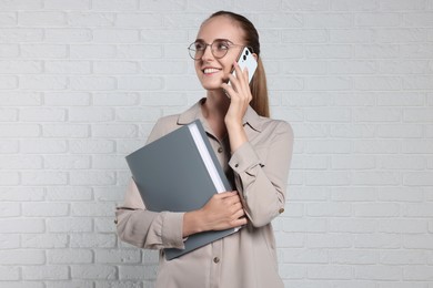 Photo of Happy young secretary with folder talking on smartphone near white brick wall