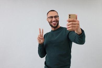 Photo of Smiling young man taking selfie with smartphone and showing peace sign on grey background, space for text