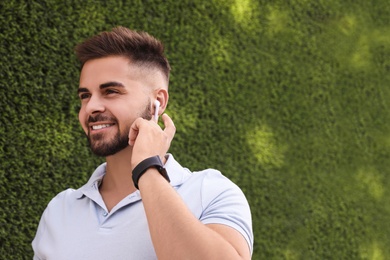 Photo of Young man with wireless headphones listening to music near green grass wall. Space for text
