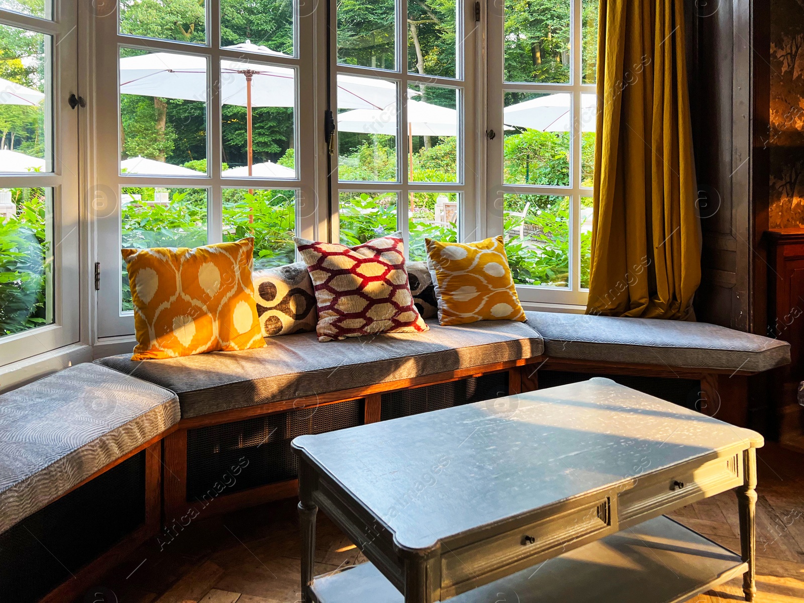 Photo of Wooden table near cozy bay window seat with cushions in room. Interior design