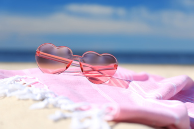 Photo of Stylish heart shaped sunglasses and blanket on sand near sea, closeup. Beach accessories for summer vacation