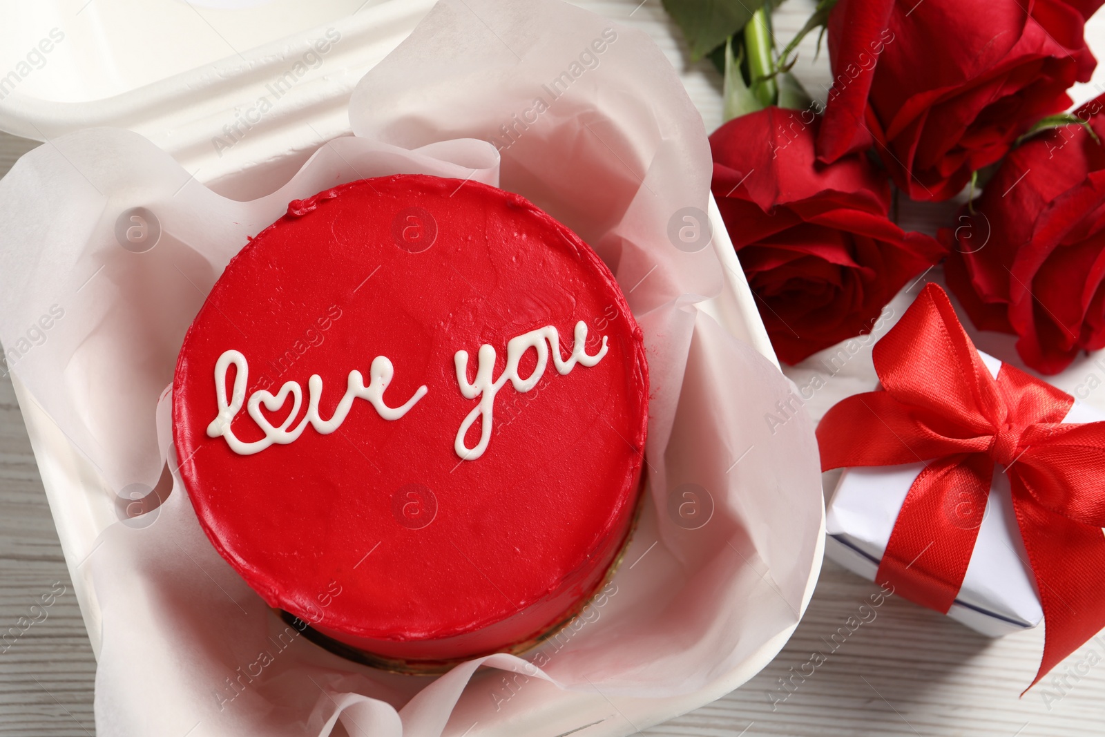 Photo of Bento cake with Love You text in takeaway packaging, roses and gift box on white wooden table, top view. St. Valentine's day surprise