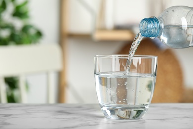 Pouring water from bottle into glass on white marble table indoors, space for text. Refreshing drink