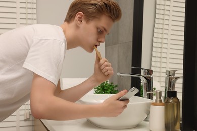 Photo of Teenage boy using smartphone while brushing teeth in bathroom. Internet addiction