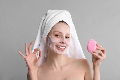 Photo of Young woman washing face with brush and cleansing foam on grey background