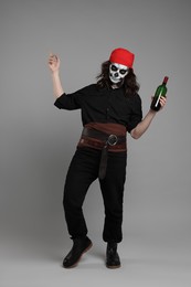 Man in scary pirate costume with skull makeup and bottle of rum on light grey background. Halloween celebration