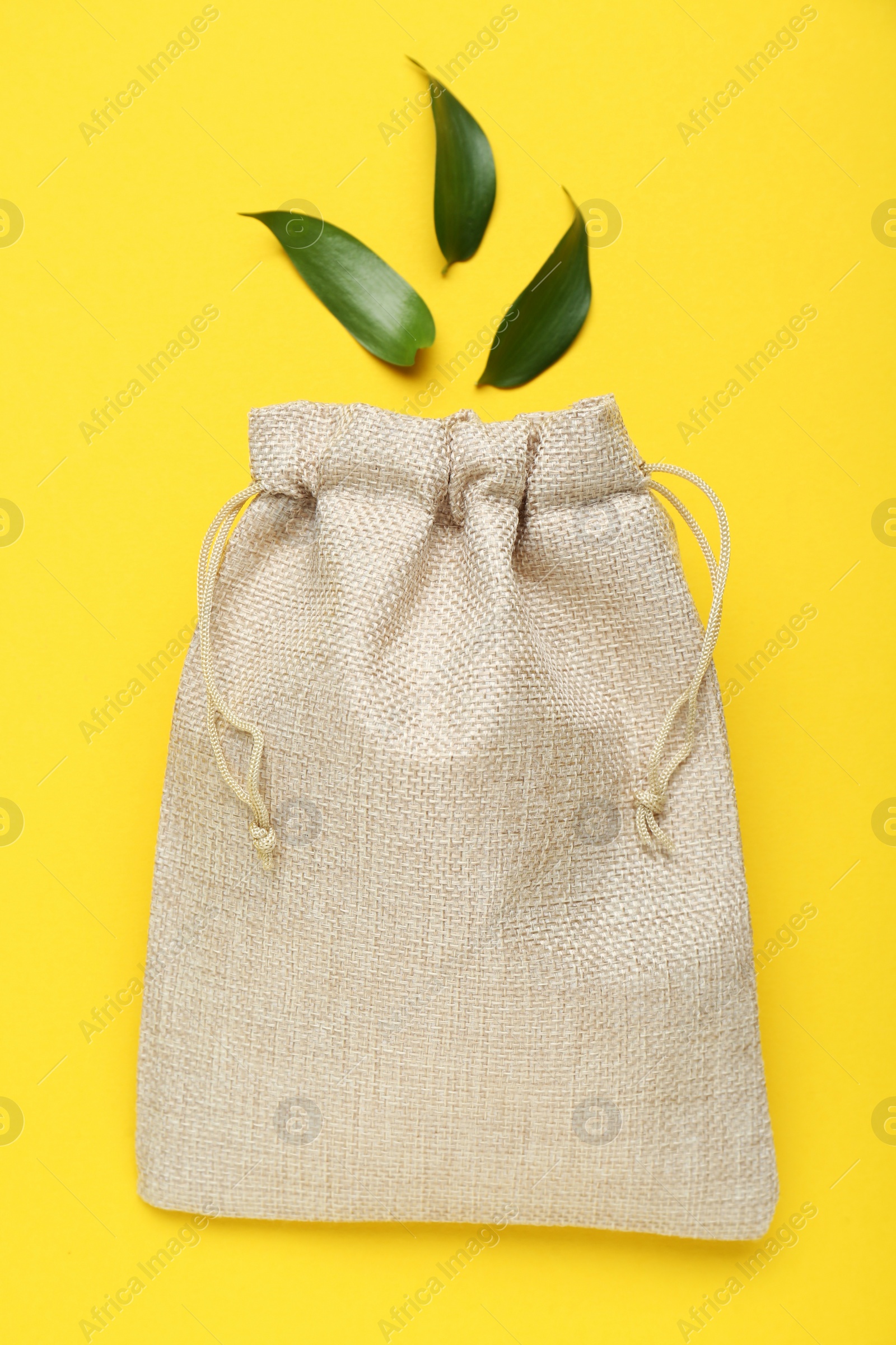 Photo of Burlap bag and green leaves on yellow background, flat lay