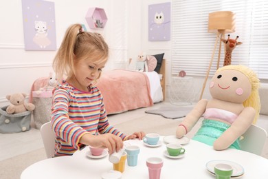 Cute little girl playing tea party with doll at table in room