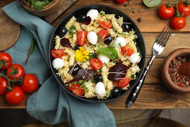 Bowl of delicious pasta with tomatoes, olives and microgreens on wooden table, flat lay