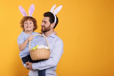 Photo of Happy father and son wearing cute bunny ears headbands on orange background. Boy holding Easter basket with painted eggs, space for text