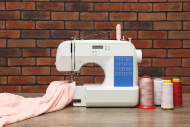 Photo of Sewing machine with fabric and spools of threads on wooden table against brick wall