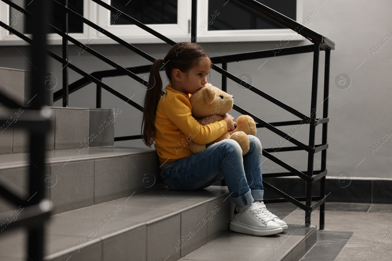 Photo of Child abuse. Upset little girl with teddy bear sitting on stairs indoors