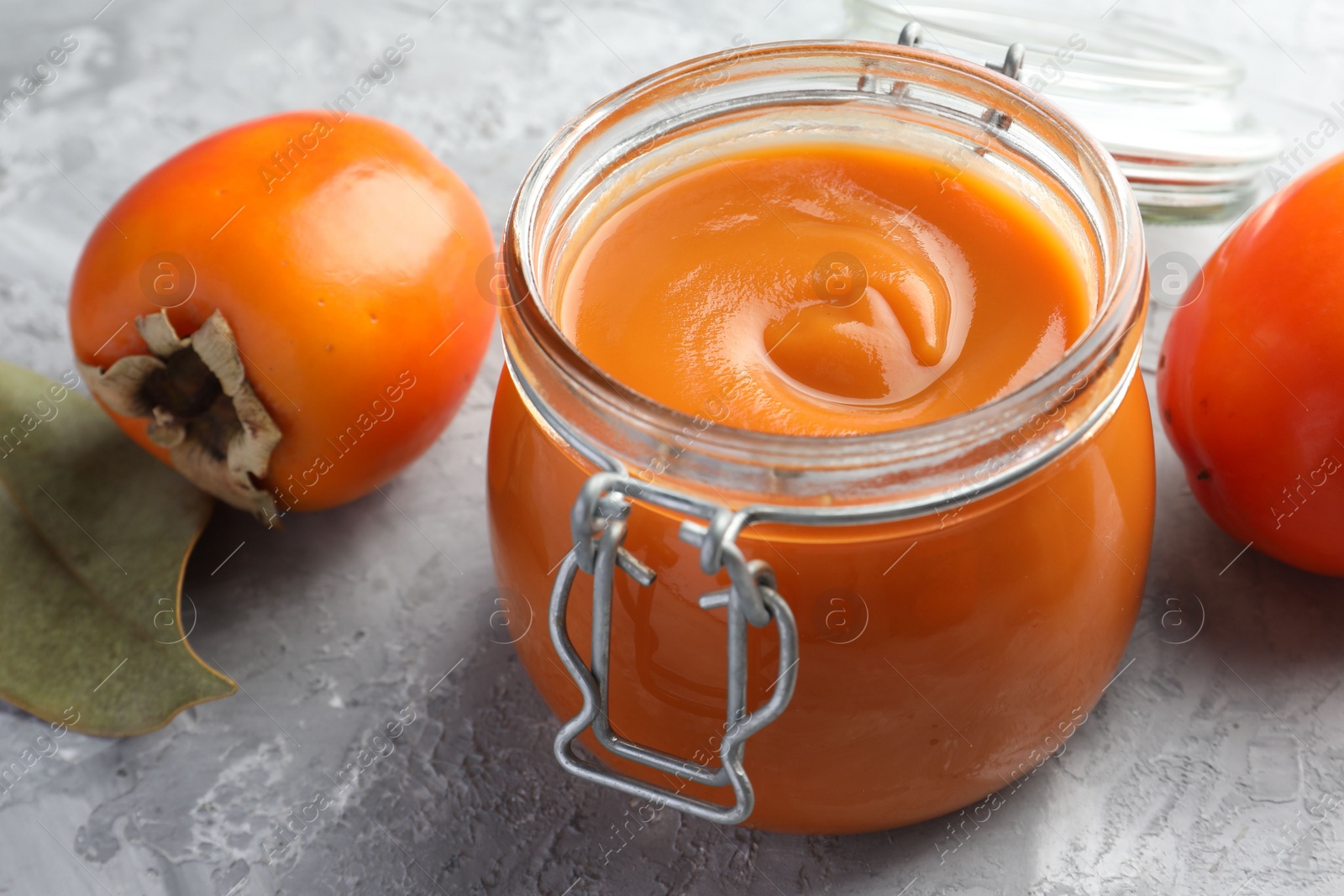 Photo of Delicious persimmon jam and fresh fruits on grey table, closeup