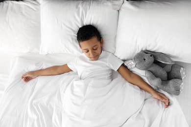 Photo of Cute little African-American boy with toy rabbit sleeping in bed, top view