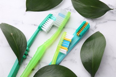 Photo of Colorful plastic toothbrushes and green leaves on white marble table, above view