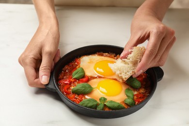 Photo of Woman dipping piece of bread into delicious Shakshuka at white table, closeup