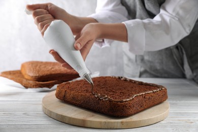Woman applying chocolate cream with pastry bag onto homemade sponge cake at white wooden table, closeup