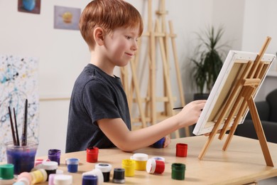 Photo of Little boy painting at table in studio. Using easel to hold canvas