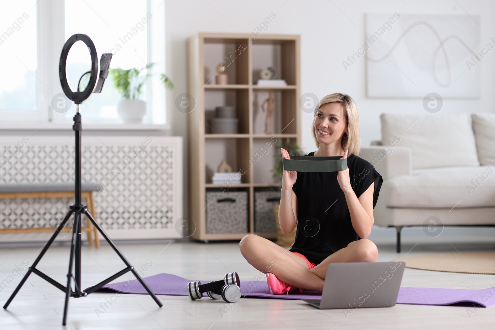 Photo of Smiling sports blogger with with resistance band streaming online fitness lesson at home
