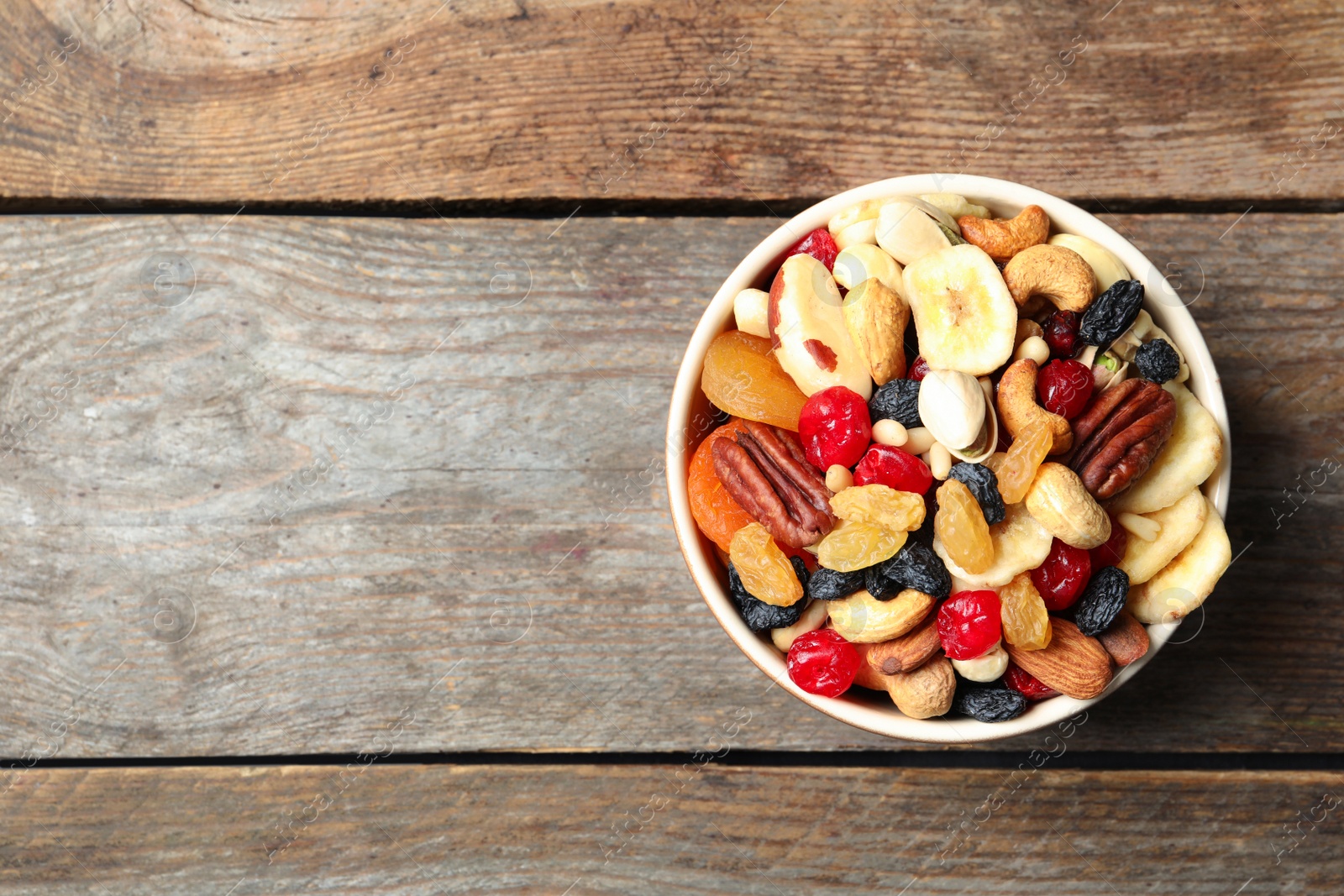 Photo of Bowl with different dried fruits and nuts on wooden background, top view. Space for text