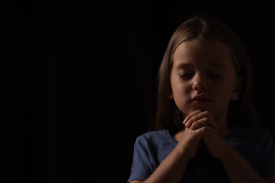 Cute little girl with hands clasped together praying on black background. Space for text